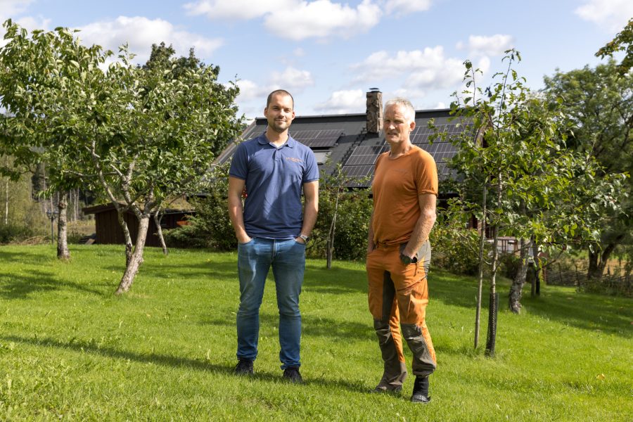 Vegard Ulvang i oransje klær og elektriker i blå arbeidsuniform står i eplehage foran hus med solceller på taket,