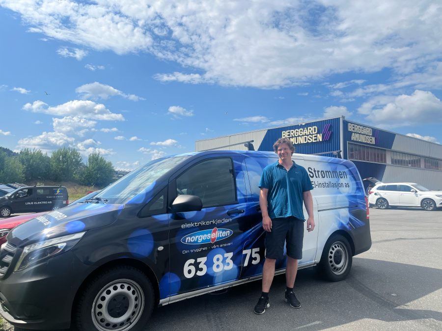 Ung mannlig elektriker står i blå shorts og t-skjorte foran firmabil parkert foran stort næringsbygg.